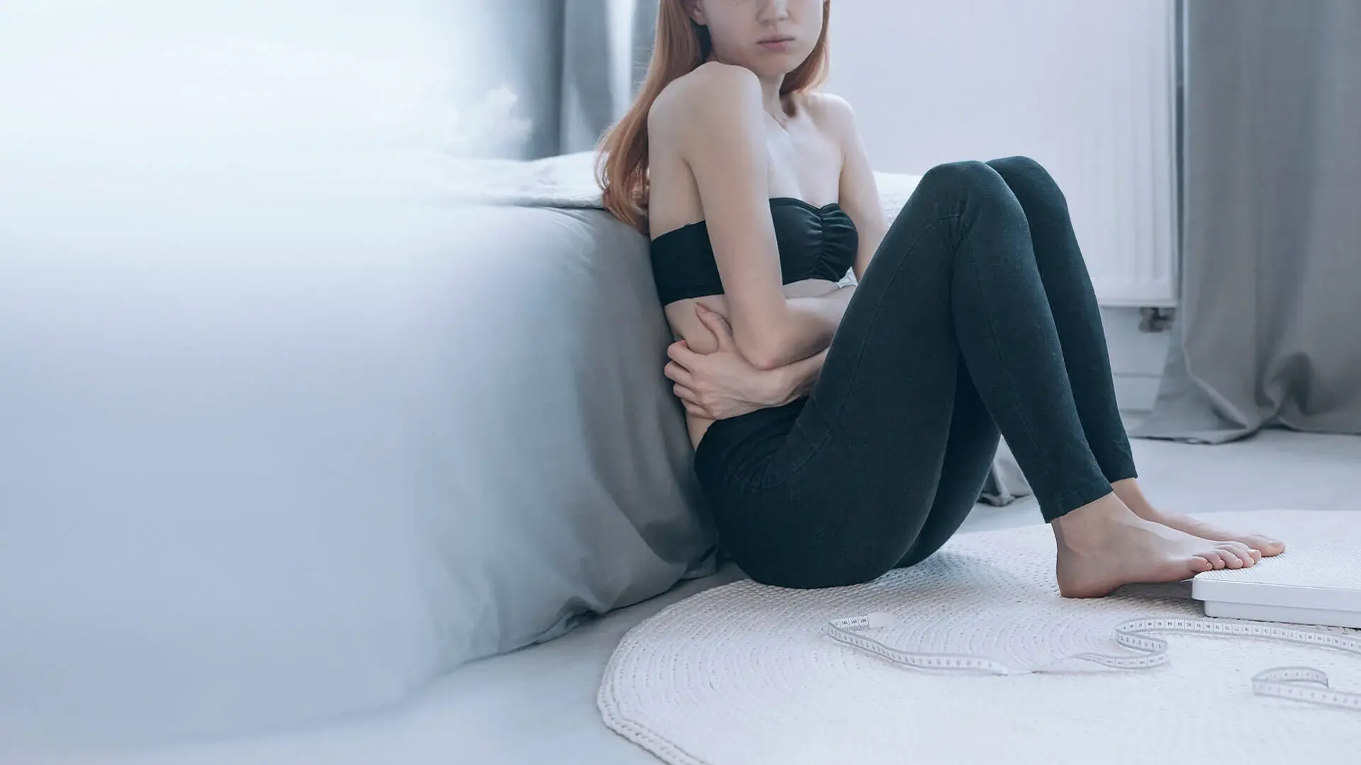 Das Symbolbild zeigt ein sehr schlankes Mädchen. Es sitzt vor dem Bett am Fußboden und hält sich den Bauch. 