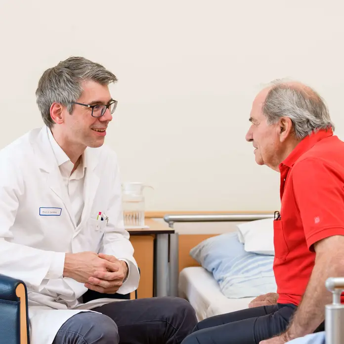 Chefarzt Prof. Dr. Alexander Dechêne spricht mit einem Patienten. Der Patient sitzt im Bett.
