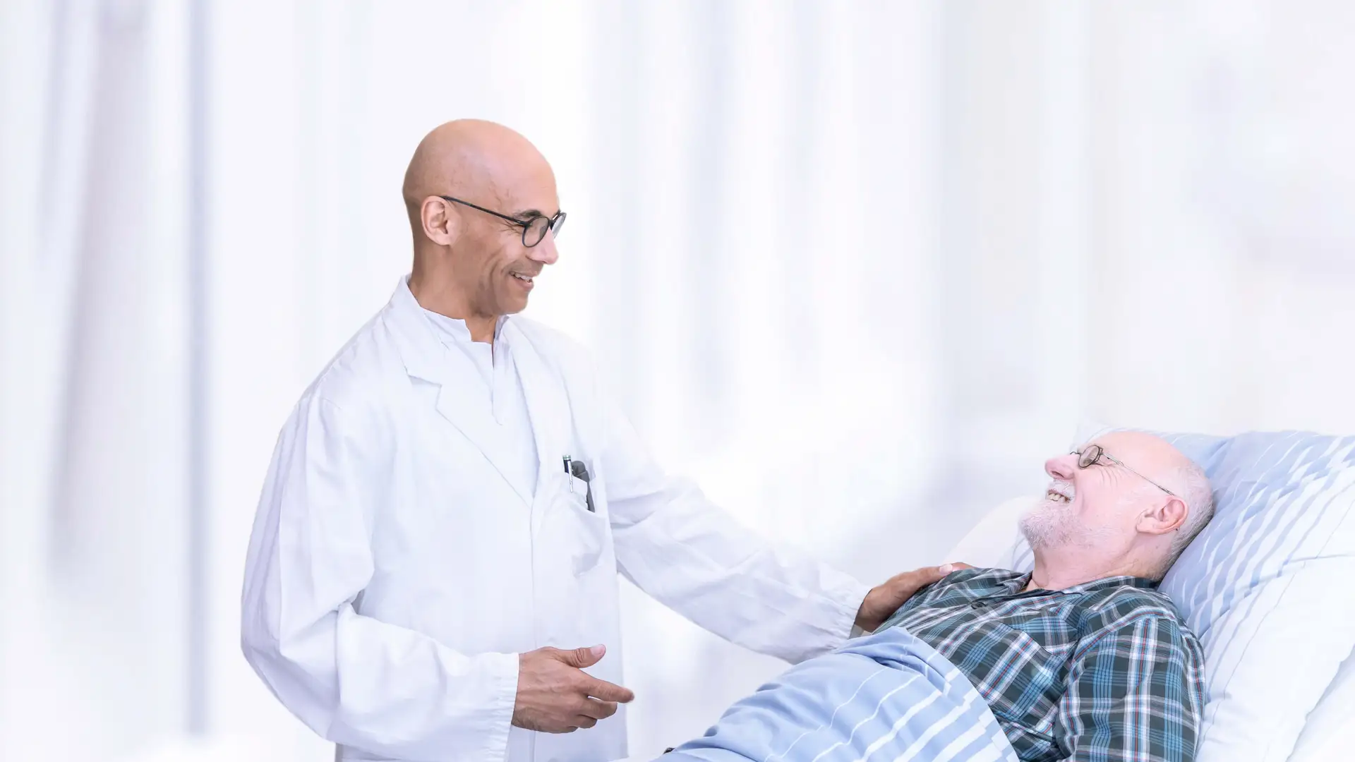 Ein Arzt spricht mit einem männlichen Patienten im Bett. Er legt seine Hand auf dessen Schulter. Die Stimmung ist positiv. 