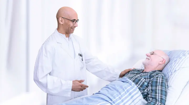 Ein Arzt spricht mit einem männlichen Patienten im Bett. Er legt seine Hand auf dessen Schulter. Die Stimmung ist positiv. 
