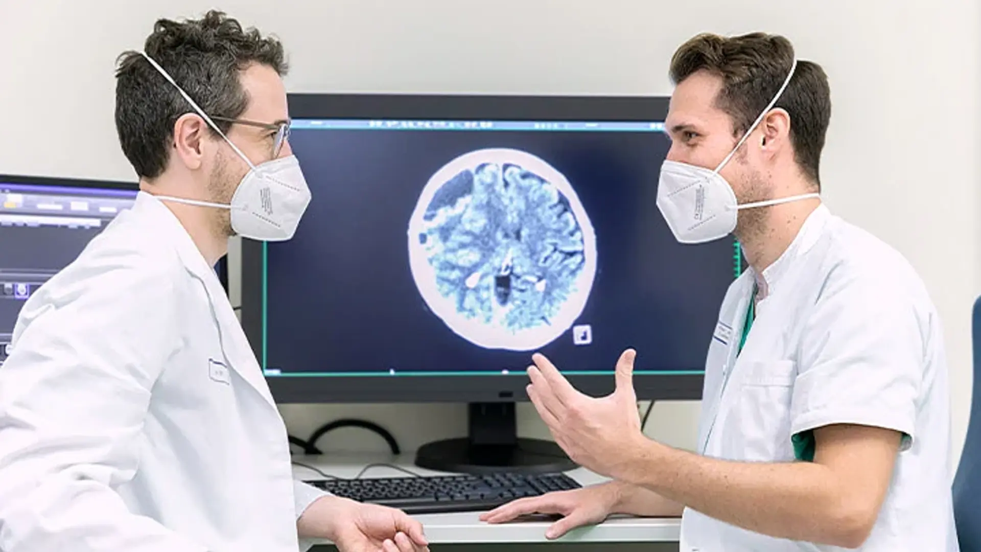 Zwei Mitarbeitende der Neuroradiologie besprechen Untersuchungsergebnisse. Sie sitzen vor mehreren Bildschirmen. 