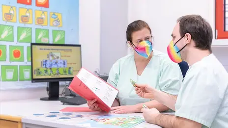 Zwei Kollegen mit bunten Masken beraten sich am Schreibtisch