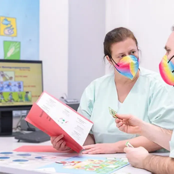 Zwei Kollegen mit bunten Masken beraten sich am Schreibtisch