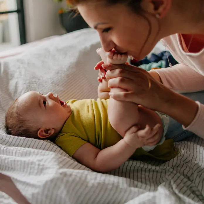 Eine junge Frau küsst die Füße eines Babys, welches auf dem Bett liegt.