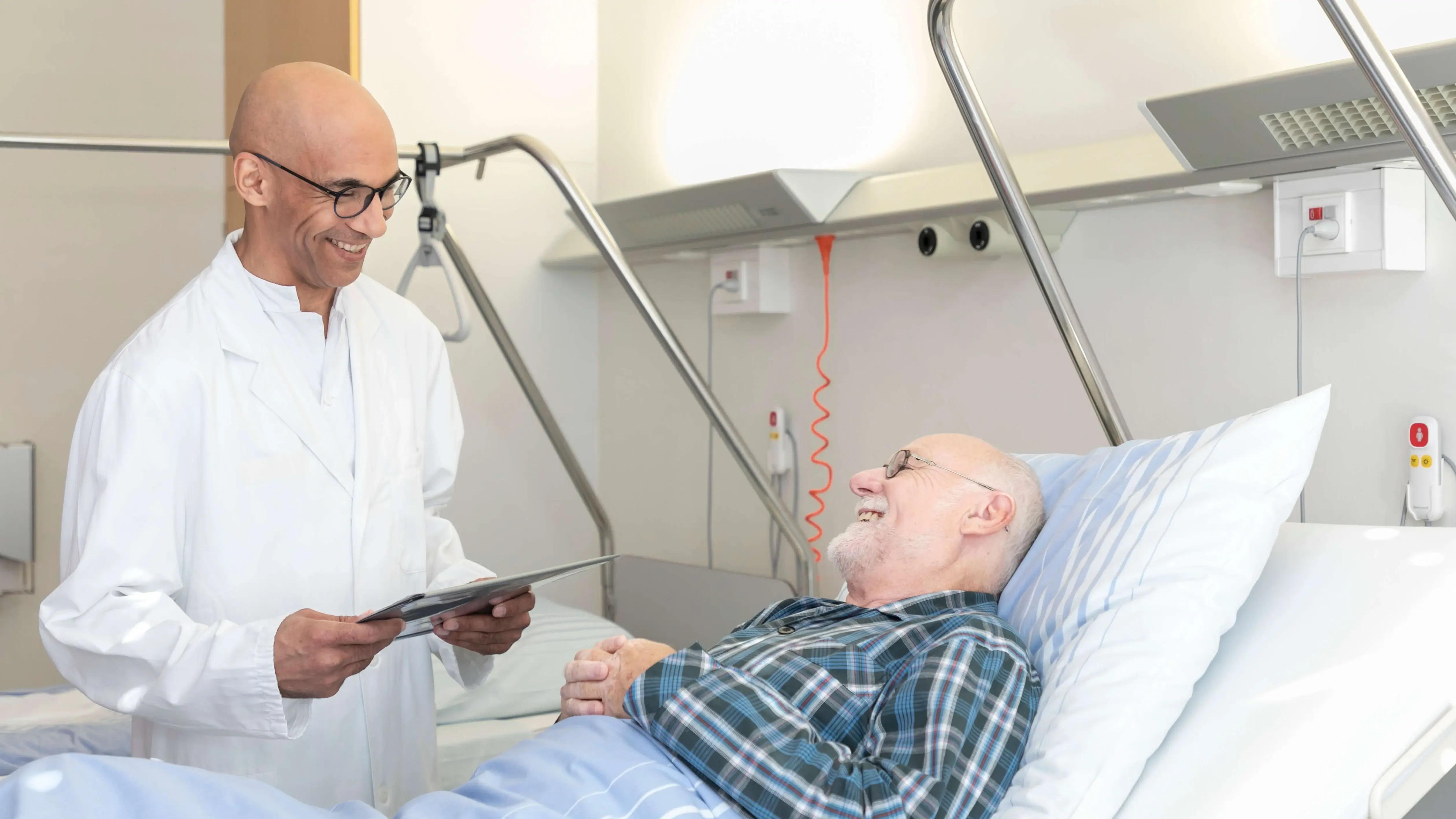 Ein Arzt spricht mit einem männlichen Patienten im Bett. Die Stimmung ist freudig. 