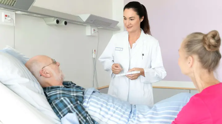 Eine Ärztin spricht mit einem Patienten im Bett. Seine Frau sitzt daneben. 