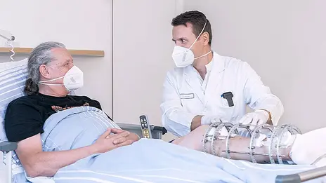 Ein Arzt der Unfallchirurgie spricht mit einem Patienten. Der Patient liegt im Bett und trägt einen Fixateur-Externe am Bein. 