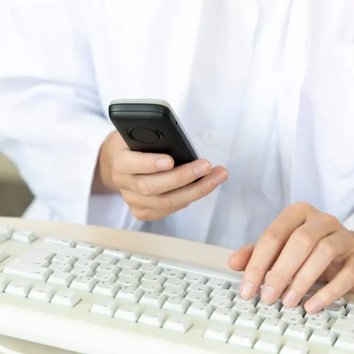 Mitarbeitende hält ein Telefon in der Hand. Die zweite Hand liegt auf der Computer-Tastatur. 