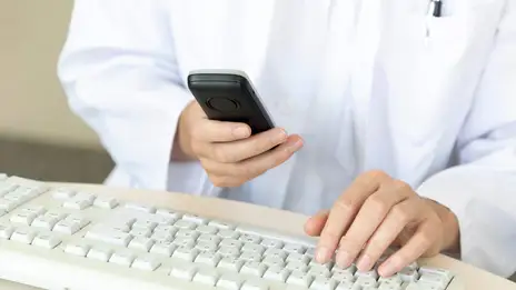 Mitarbeitende hält ein Telefon in der Hand. Die zweite Hand liegt auf der Computer-Tastatur. 