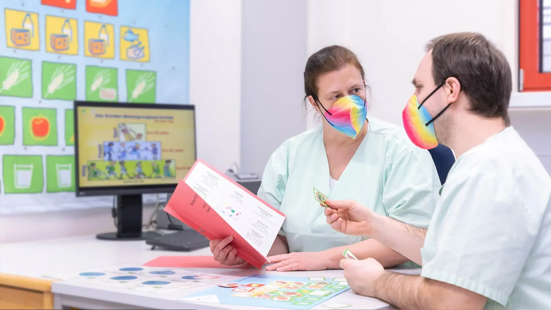 Zwei Kollegen mit bunten Masken beraten sich am Schreibtisch
