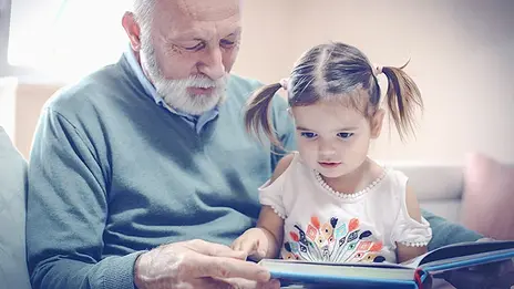 Das Symbolbild zeigt einen älteren Herren, der einem Mädchen ein Buch vorliest. 
