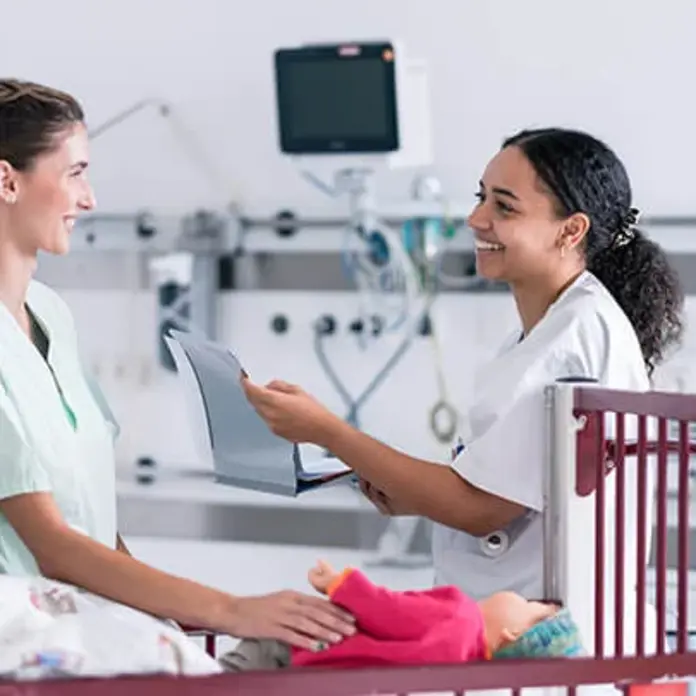 Die zwei Pflegekräfte schauen sich in die Augen. Die Frauen stehen vor einem Kinderbett. 