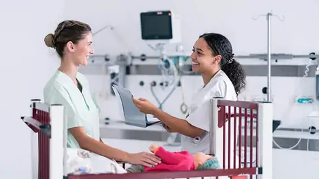Die zwei Pflegekräfte schauen sich in die Augen. Die Frauen stehen vor einem Kinderbett. 
