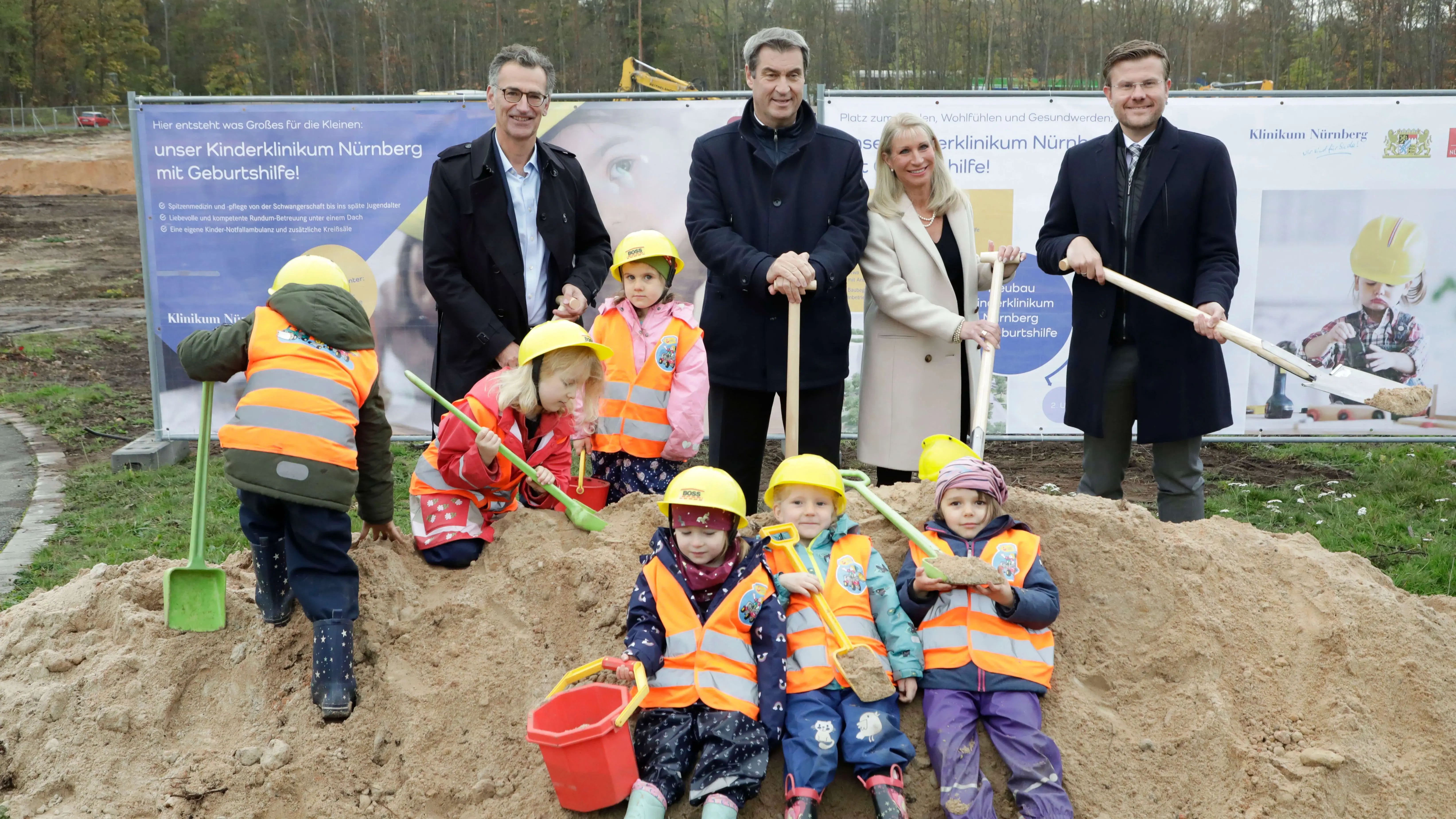 Gruppenbild vor einem symbolischen Erdhaufen. Zu sehen sind Ministerpräsident Dr. Markus Söder, Oberbürgermeister Marcus König, Schirmherrin Karin Baumüller-Söder und Klinikums-Vorstand Prof. Dr. Achim Jockwig. Auf dem Erdhügel sitzen Kinder mit Warnwesten und gelben Schutzhelmen. 