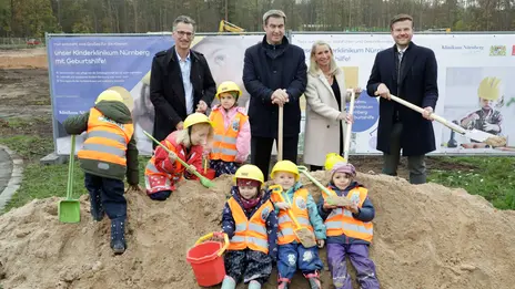 Gruppenbild vor einem symbolischen Erdhaufen. Zu sehen sind Ministerpräsident Dr. Markus Söder, Oberbürgermeister Marcus König, Schirmherrin Karin Baumüller-Söder und Klinikums-Vorstand Prof. Dr. Achim Jockwig. Auf dem Erdhügel sitzen Kinder mit Warnwesten und gelben Schutzhelmen. 