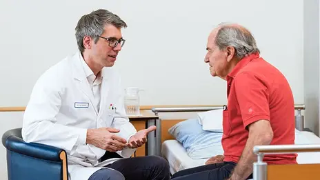 Chefarzt Prof. Dr. Dechêne spricht mit einem Patienten. Der Patient sitzt in seinem Bett. 