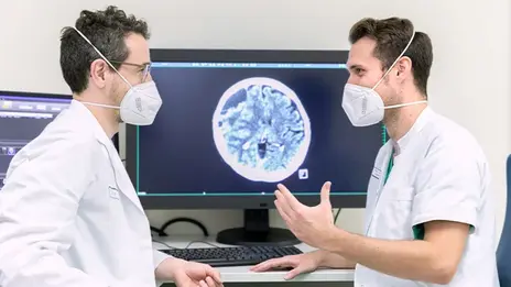 Zwei Mitarbeitende der Neuroradiologie besprechen Untersuchungsergebnisse. Sie sitzen vor mehreren Bildschirmen. 
