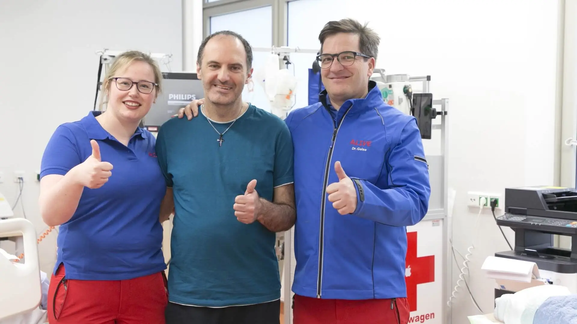 Zwei Mitarbeitende des Klinikums und der gerettete Patient lachen in die Kamera. 