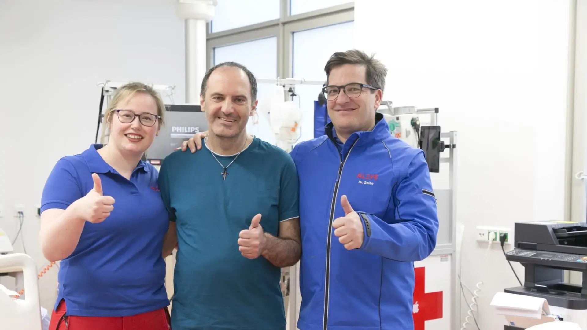 Zwei Mitarbeitende des Klinikums und der gerettete Patient lachen in die Kamera. 