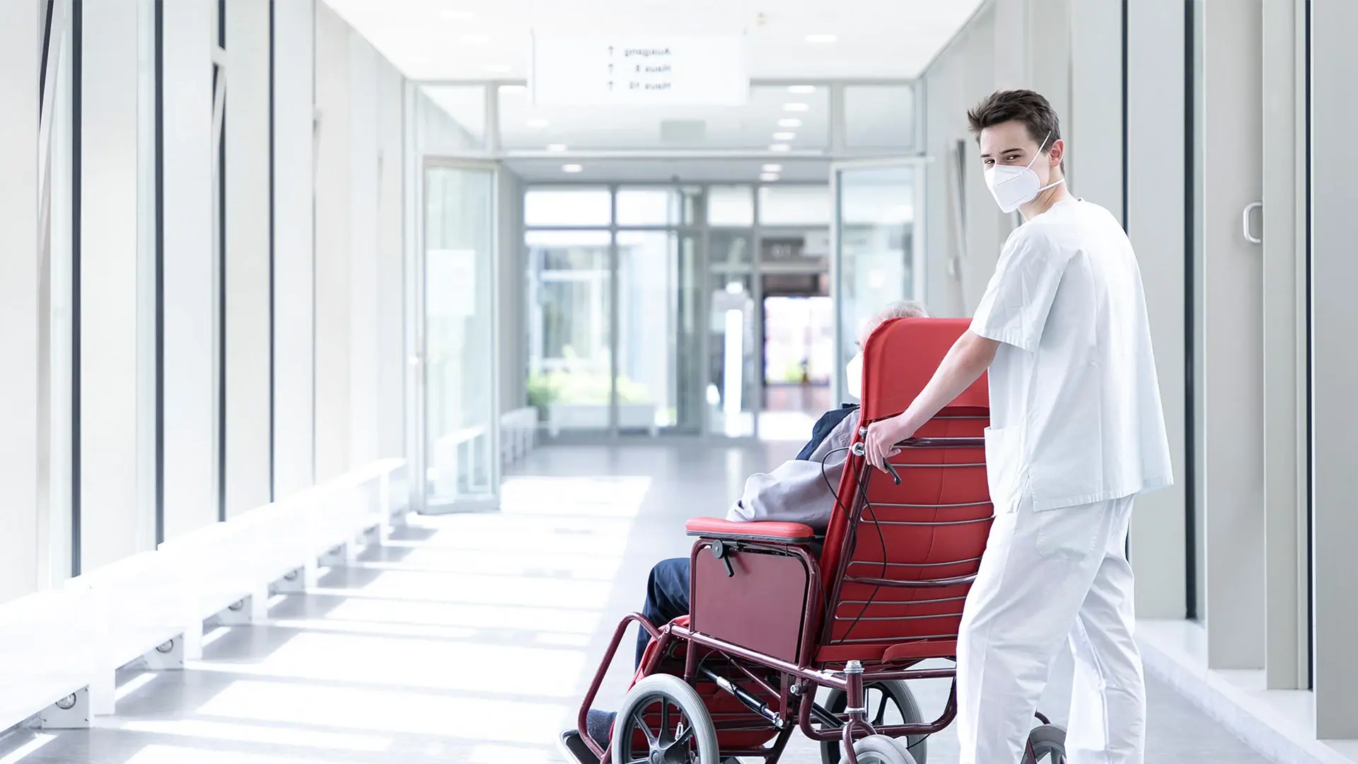 Ein Mitarbeiter schiebt einen Patienten im Rollstuhl zur nächsten Untersuchung. 