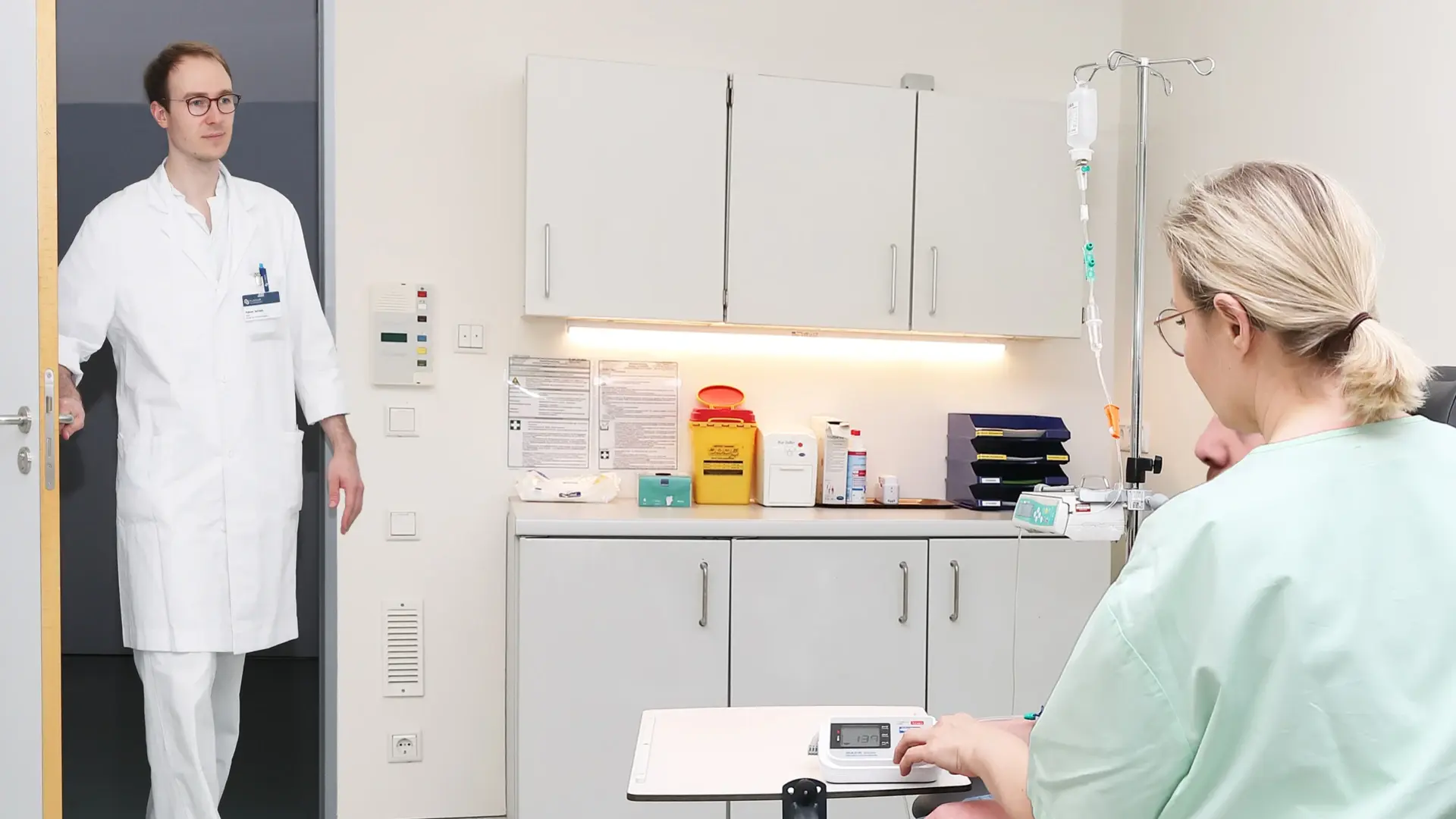 Ein Arzt betritt das Behandlungszimmer wo die Pflegefachkraft mit dem Patienten bereits auf ihn warten.