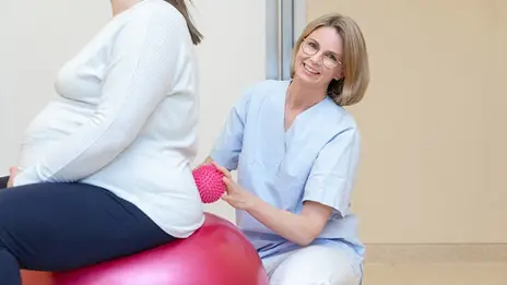 Eine Hebamme stützt eine schwangere Frau auf dem Gymnastikball.