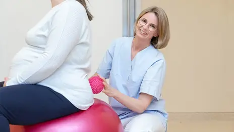 Eine Hebamme stützt eine schwangere Frau auf dem Gymnastikball.